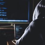 hacker in hoodie typing code in program using laptop in dark studio